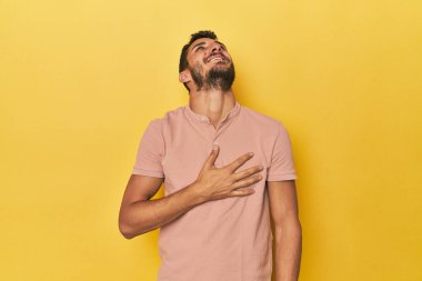 Sarı arka plandaki genç İspanyol adam elini göğsünde tutarak kahkahalar atıyor..