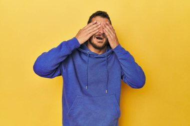 Sarı arka plandaki genç İspanyol adam elleriyle gözlerini kapatıyor. Gülümsüyor ve bir sürpriz bekliyor..