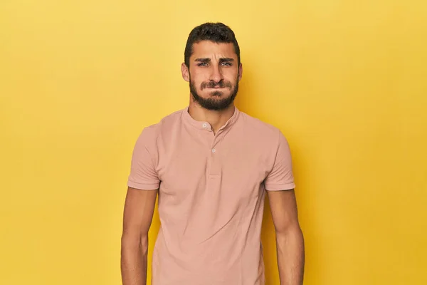 Young Hispanic man on yellow background blows cheeks, has tired expression. Facial expression concept.