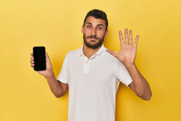 年轻的拉美裔男子在手机屏幕上开心地微笑着 用手指显示第五位 图库图片