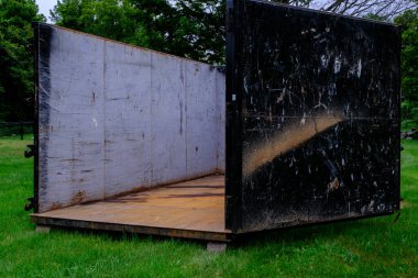 İyi kullanılmış, siyah ve gümüş renkli endüstriyel bir çöp konteynırında toprak, sürtünme ve daha önce kullanıldığına dair izler var. Arka planda ağaçlar olan yeşil çimlerin üzerine yerleştirilmiş. Yenileme enkazı ve inşaat malzemeleri taşımak için ideal..