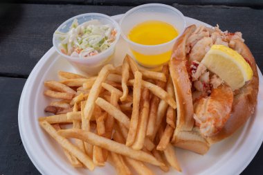 Patates kızartması, lahana salatası ve tek kullanımlık beyaz bir tabakta erimiş tereyağı ile servis edilen taze bir ıstakoz rulosu. Klasik bir New England deniz ürünleri yemeği..