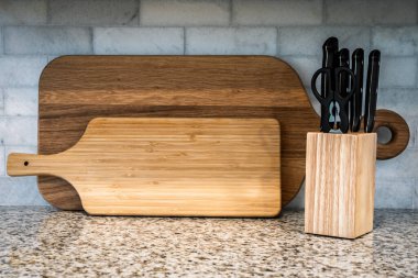 Temiz ve modern mutfak sahnesinde iki ahşap kesme tahtası ve siyah saplı bıçaklar ve mutfak makası olan bir bıçak bloğu, taş kaplama ve granit tezgaha karşı dizilmiş. Yemek pişirme, aşçılık ya da ev hayatı temaları için ideal.