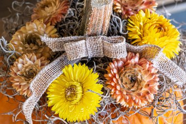 Close-up of a rustic autumn decoration featuring an orange pumpkin adorned with dried flowers, burlap ribbon, and natural moss. The bright yellow and peach flowers add a touch of seasonal color and texture.