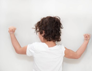 Yeni yürümeye başlayan bir çocuk kaslarını kasarak küçük pazılarını ve güçlü kollarını beyaz bir duvara dayar. Dirseği bükülmüş, başparmağı havada, yüzünde şakacı bir ifade var.