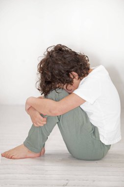 Genç bir çocuk ahşap zeminde oturuyor, başı eğik, dizleri ve kalçaları yerde dinleniyor. Başparmağı koluyla kıpırdıyor, basit bir hareketle teselli arıyor.