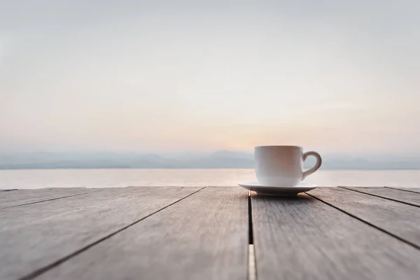 Cup of coffee on wooden deck over sunrise with river mountain in background
