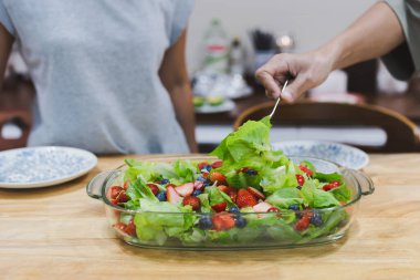Elinde çatalla taze sebze ve meyve salatasını tabağa koyuyor.
