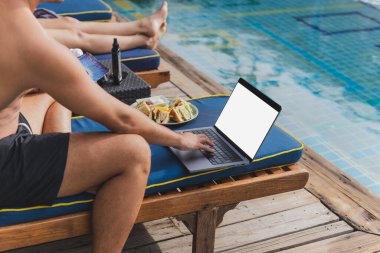 Tatilde dizüstü bilgisayarda çalışan adam havuz başında kulüp sandviçi yiyor.