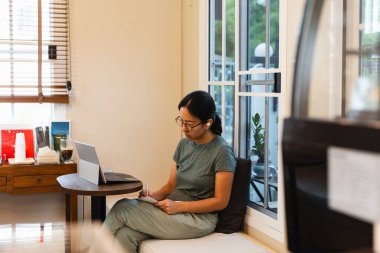 Dizüstü bilgisayar üzerinde çalışırken tükenmez kalemle not alan kadın