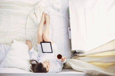 Sabahları yatakta otururken tablet kullanan ve kahve içen kadın portresi.