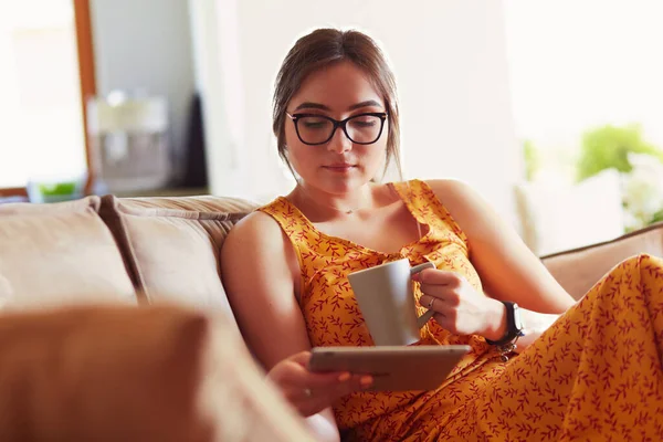 年轻女性在沙发上喝咖啡的同时 还经常使用平板电脑 — 图库照片