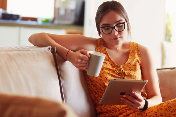 年轻女性在沙发上喝咖啡的同时 还经常使用平板电脑 — 图库照片