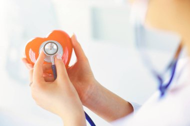 Steteskoplu bir doktor, beyaz arka planda kırmızı kalbi inceliyor..