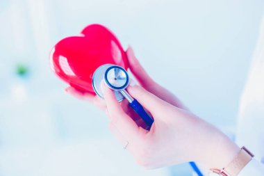 Steteskoplu bir doktor, beyaz arka planda kırmızı kalbi inceliyor..