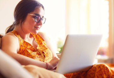 Evde dizüstü bilgisayar üzerinde çalışan kanepede oturan genç kadın.