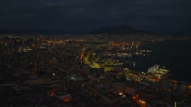 İleri uçaklar gece şehir merkezinin üzerinde uçar. Aydınlatılmış sokaklar ve deniz kıyısındaki şehir limanı. Vezüv Dağı silueti uzakta. Napoli, İtalya.
