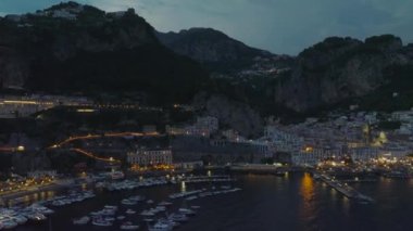 Deniz kıyısındaki teknelerin üzerinde ilerleyin. Şehirdeki tarihi binalar ve arka planda yüksek kayalar. Akşamları turistik bir yerde. Amalfi, İtalya.