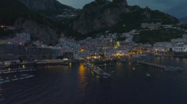 Eski kasabadaki deniz kıyısının havadan görünüşü. Dalgalı su yüzeyi ışıkları yansıtıyor. Geriye doğru demirli teknelerdeki sıraları ortaya çıkarıyor. Amalfi, İtalya.