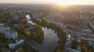 Gün batımında şehir merkezinden akan nehrin resimli hava görüntüleri. Kıyılarda sonbahar ağaçlarıyla çevrili sakin su yüzeyinde yelken açan bir tekne. Berlin, Almanya.
