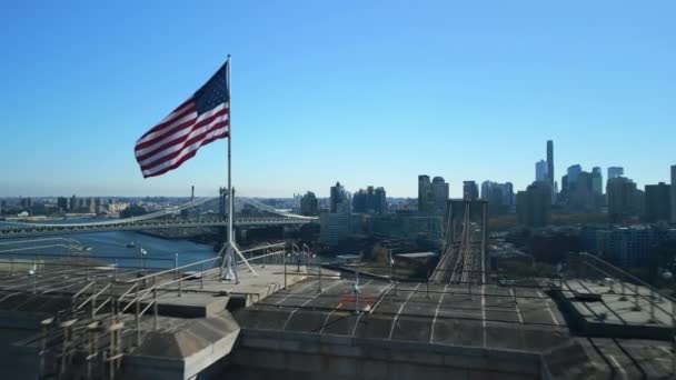 前進はブルックリン橋の上で風になびく米国の旗の周りを飛ぶ 都市のバラの橋や建物のパノラマビュー 米国ニューヨーク市 — ストック動画