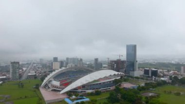 Aerial cinematic shot of Estadio Nacional. Modern multipurpose sports venue surrounded by park and high rise buildings in city. San Jose, Costa Rica.