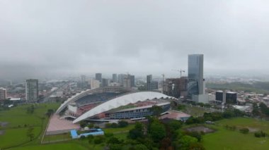 Modern multipurpose stadium in La Sabana Metropolitan Park and high rise town development in urban borough. Aerial view of Estadio Nacional on hazy day. San Jose, Costa Rica.