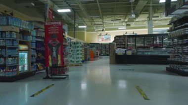 Low flight between shelves with products in shop. Scene lit by dimmed lights. Interior of large supermarket outside opening hours.