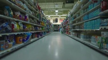 Low flight in shop aisle surrounded by multilevel shelves with arranged goods. Interior of large supermarket outside opening hours.