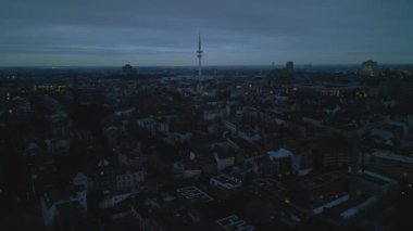 Forwards fly above buildings in residential urban borough at dusk. Heading towards tall TV tower. Hamburg, Germany.