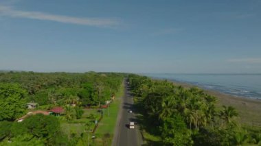 Aerial view of asphalt road sneaking through rainforest near beach. Flying over an expressway into tropical lush greenland near exotic white sand beach.