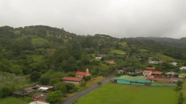 Aerial slide and pan footage of group of buildings in small town in tropical landscape. Chapel with tower at road. La Fortuna, Costa Rica.