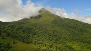 Aerial ascending footage of steep slope of hill overgrown with green vegetation. Majestic peak of Arenal volcano, clouds moving around top. La Fortuna, Costa Rica.