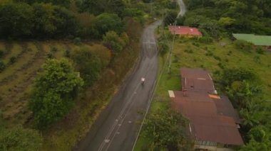 Forwards tracking of biker passing through village on motorbike. Aerial view of hilly landscape at rain. La Fortuna, Costa Rica.