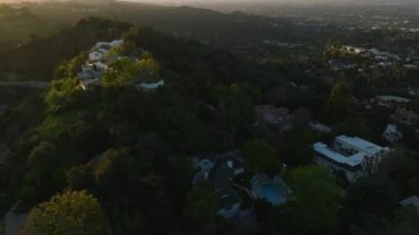 Şehrin tepelerindeki ağaçlarla çevrili lüks evlerin üzerinde uç. Gün batımında şehir manzarası. Los Angeles, Kaliforniya, ABD.
