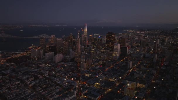 日落后商业中心大楼的空中电影镜头 灯光明亮的摩天大楼和背景在海湾上方的桥 美国加利福尼亚州旧金山 — 图库视频影像
