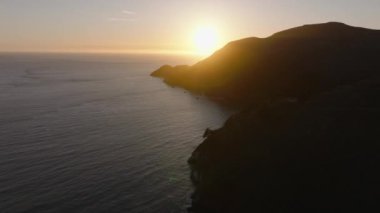 Batan güneşe karşı okyanus kıyısında ilerliyorlar. Sahildeki tepelerin silueti. San Francisco, California, ABD.