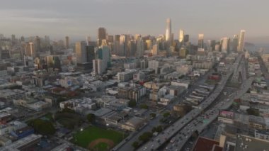 Şehir merkezindeki gökdelenlerin güneşin batışıyla aydınlanan panoramik görüntüleri. Çok şeritli anayolda trafik sıkışık. San Francisco, California, ABD