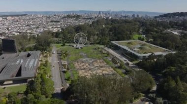 İleri, Golden Gate Parkı 'nın üzerinden uçar. Yeşil çimenler ve ağaçlarla çevrili anıtlar ve müze binaları. Şehrin arka planında binalar var. San Francisco, California, ABD.