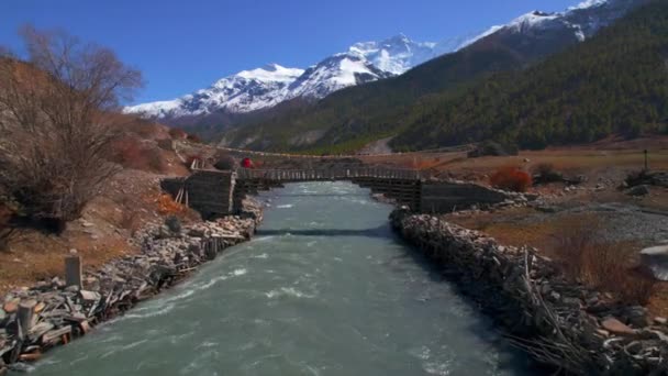 ハイカーは野生の川を渡る木製の橋の上を歩いています バックグラウンドのピークに雪が降る山の風景 マナンバレー アンナプルサーキットトレック ネパール — ストック動画