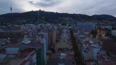 Şehir merkezindeki çok katlı binaların üstünde alçak uçuş. TV kulesinin silueti ve tepedeki kilise alacakaranlık gökyüzüne karşı. Barselona, İspanya.