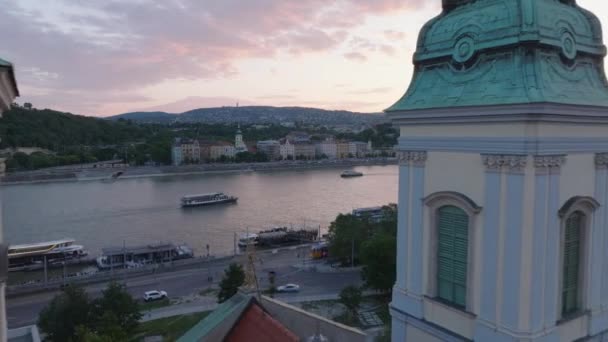 2つの教会の塔の間を飛行する ウォーターフロントを通過するトラムと広い川を巡航する観光船を明らかにします ブダペスト ハンガリー — ストック動画