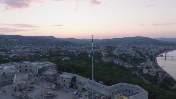 オービットはハンガリーの国旗の周りを 市の上の丘の上の要塞のポールで撃ちました トワイライトの大都市の空中パノラマビュー ブダペスト ハンガリー — ストック動画