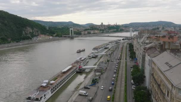 フォワードは 曇りの日に市内の広いダヌーブ川のウォーターフロントの上を飛行します 銀行に沿った大きな観光船 ブダペスト ハンガリー — ストック動画