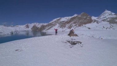 Hava dairesel manzaralı erkek yürüyüşçü zirvede duruyor. Kollarını kaldırarak trekking hedefi ve hedefine ulaşmanın keyfini çıkarıyor. Açık hava macerası, Himalaya, Asya, Tilicho 'da yürüyüş..