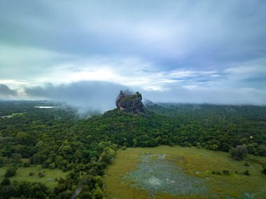 Çevredeki ormanın üzerinde yükselen antik kaya kalesinin havadan görünüşü. Ormanın üzerindeki alçak bulutlar. Sigirya, Sri Lanka.