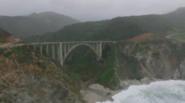 Big Sur kıyısındaki Bixby Creek Köprüsü 'nden geçen arabaların hava görüntüsü. Büyük kemerli yüksek beton köprü. Uçurumun altındaki kumsalı dalgalandırır. Kaliforniya, ABD.