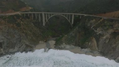 Deniz kıyısındaki Bixby Creek Köprüsü 'nün hava görüntüleri. Dalgalar kumda yuvarlanıyor. Arka plandaki dağ manzarasının puslu görüntüsü. Kaliforniya, ABD.
