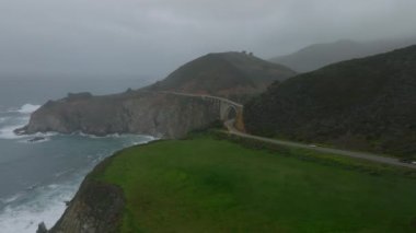 Deniz kıyısı boyunca kıvrılan panoramik manzaranın hava görüntüsü. Güzel, yüksek kemerli bir köprü ortaya çıkıyor. Kaliforniya, ABD.