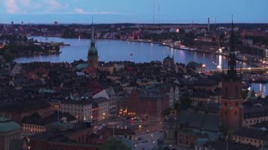 Yüksek kilise kuleleri ve popüler turizm manzaralarıyla tarihi şehir merkezinin üzerinden uç. Su yüzeyi ve şehir akşam karanlığında. Stockholm, İsveç.